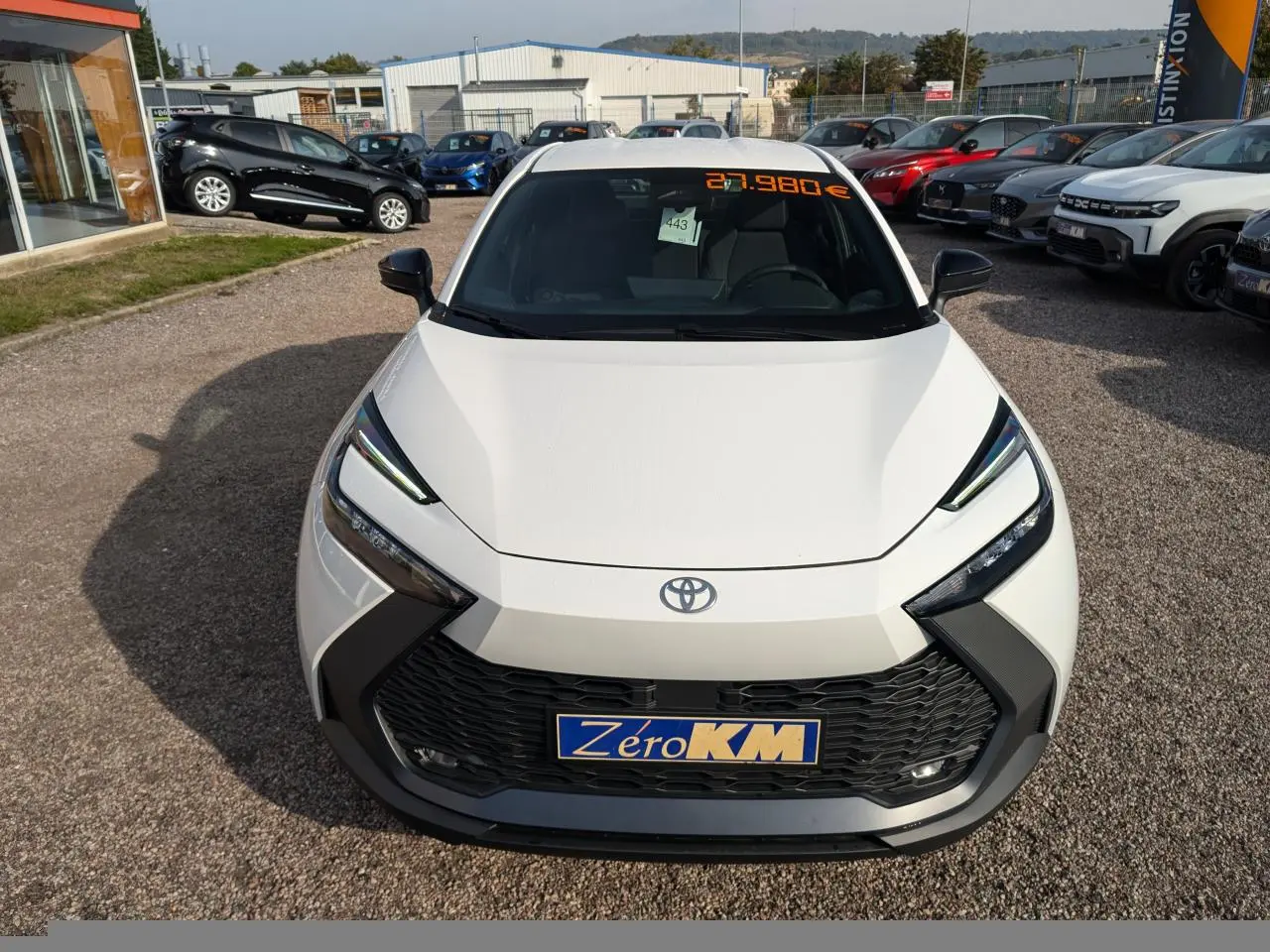 Vue avant d'un Toyota C-HR blanc 2024 avec calandre noire et feux LED, affichage prix sur pare-brise.