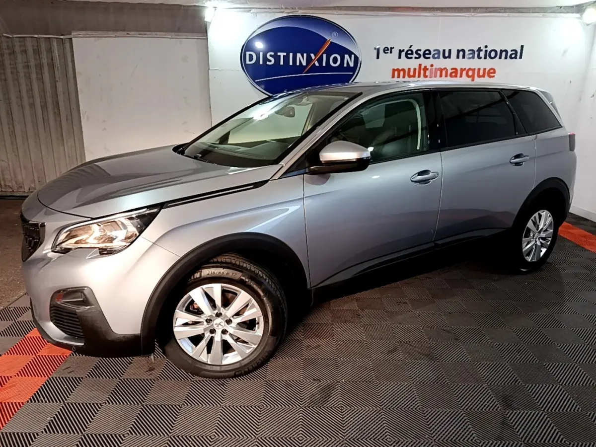 Peugeot 5008 gris clair vue 3/4 avant droit en intérieur, avec jantes alliage et feux allumés.