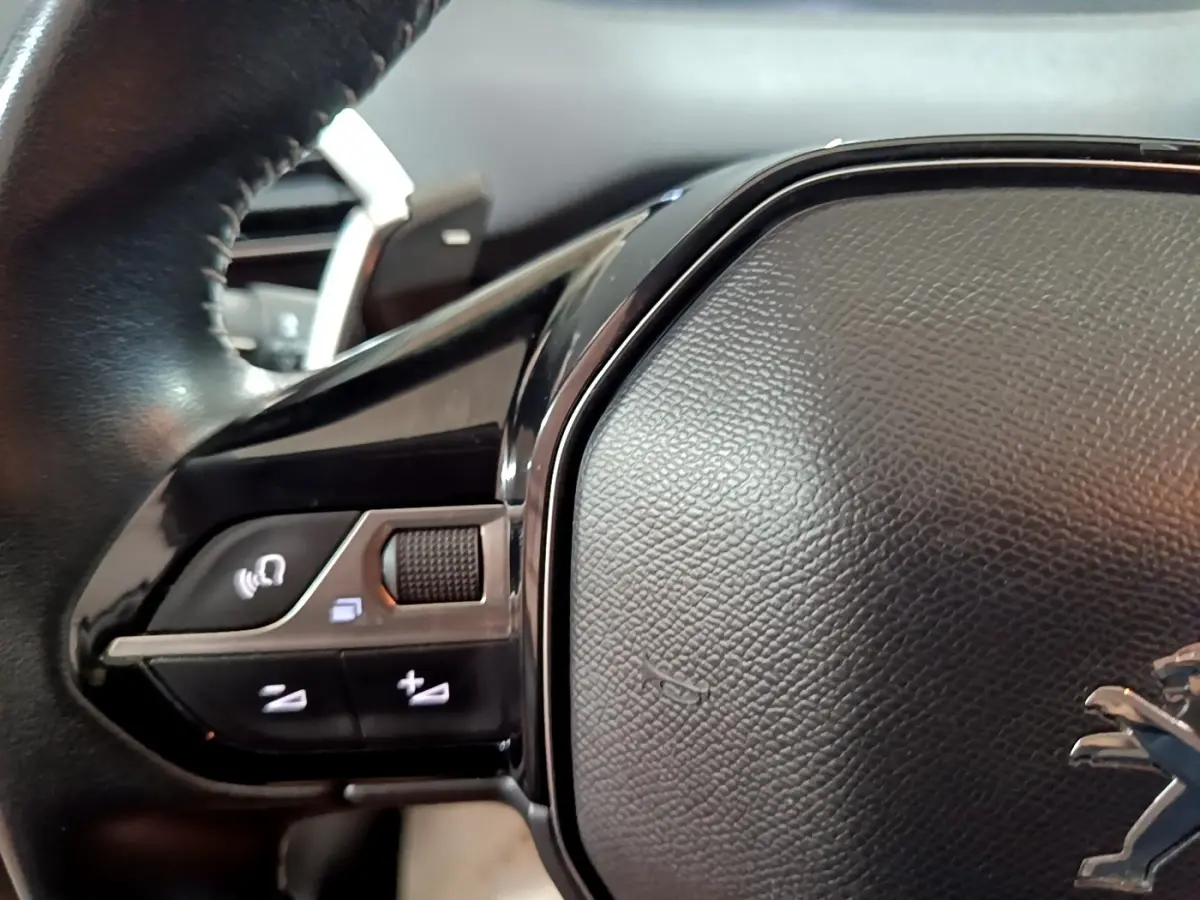 Gros plan sur le volant cuir noir du Peugeot 5008 gris clair, avec commandes multifonctions et logo lion visible.