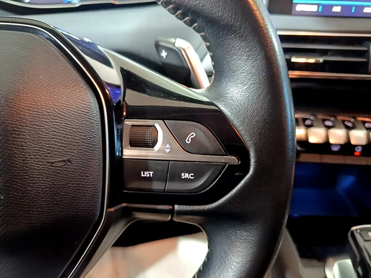 Gros plan sur la partie gauche du volant cuir noir du Peugeot 5008, avec commandes audio et téléphone intégrées.