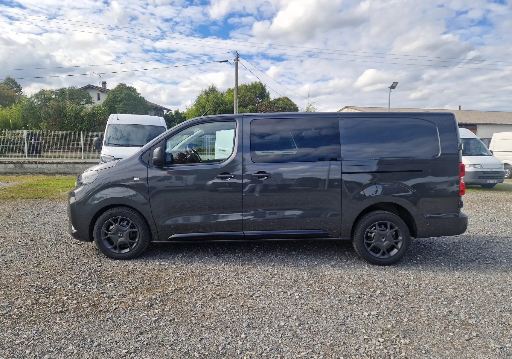 Profil côté gauche d'un Citroën Jumpy Cabine Approfondie gris titane avec jantes tôles noires sur un sol gravillonné.