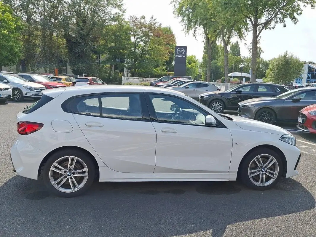 Profil côté gauche d'une BMW Série 1 blanche 120i M Sport 2021, avec jantes aluminium et lignes dynamiques visibles.