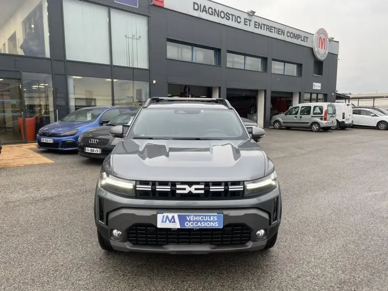 Vue avant d'un Dacia Duster 1.6 Hybrid gris schiste 2025 avec calandre distinctive et toit équipé de barres de toit.