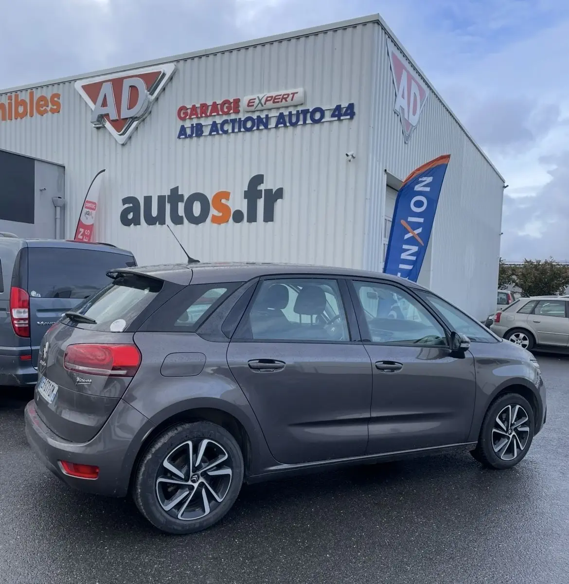 Vue côté gauche arrière d'une Citroën C4 Picasso marron avec jantes alu, stationnée devant un garage autos.fr par temps couvert.