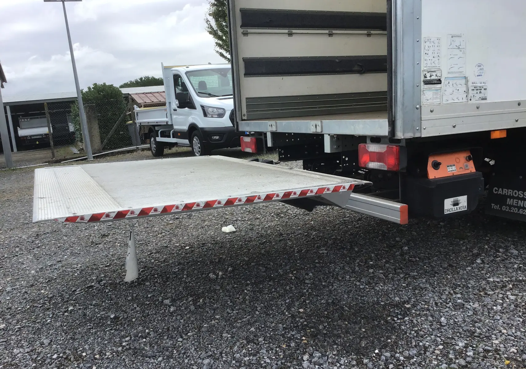 Vue arrière d'un Iveco Daily blanc avec hayon élévateur déployé sur un sol gravillonné, en extérieur.