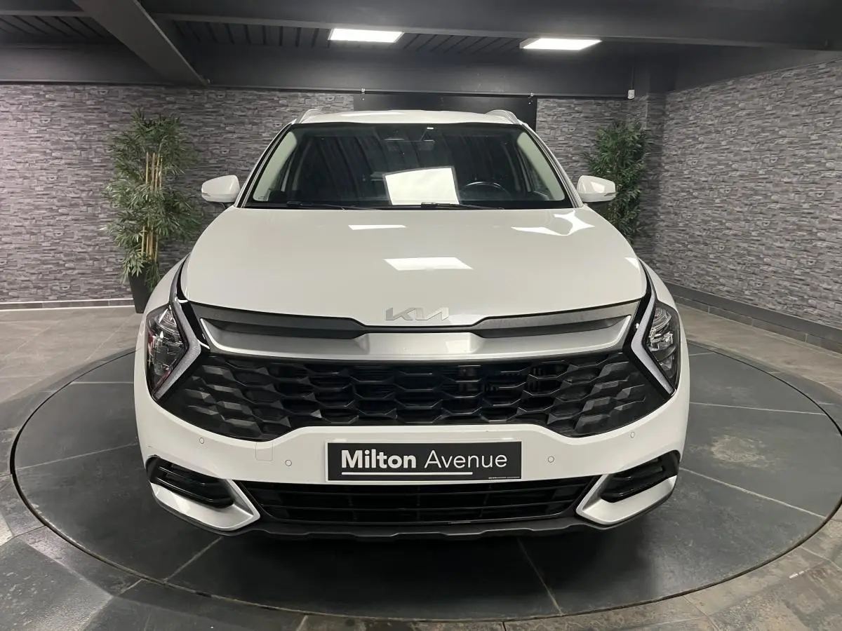 Vue frontale d'un KIA Sportage blanc nacré avec calandre noire laquée et éclairage LED en intérieur showroom.