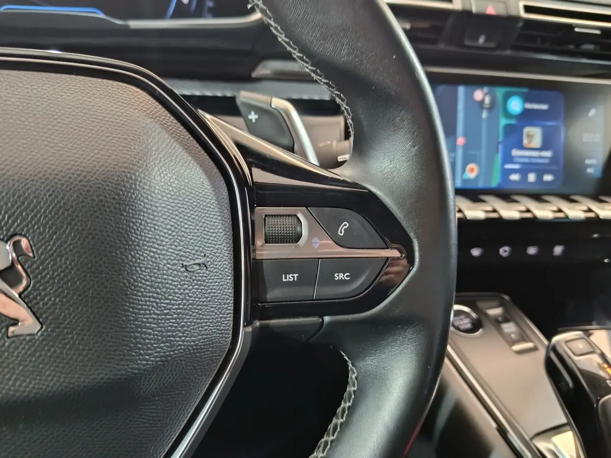 Gros plan sur le volant cuir côté droit de la Peugeot 508 gris clair, avec commandes audio et écran tactile en arrière-plan.