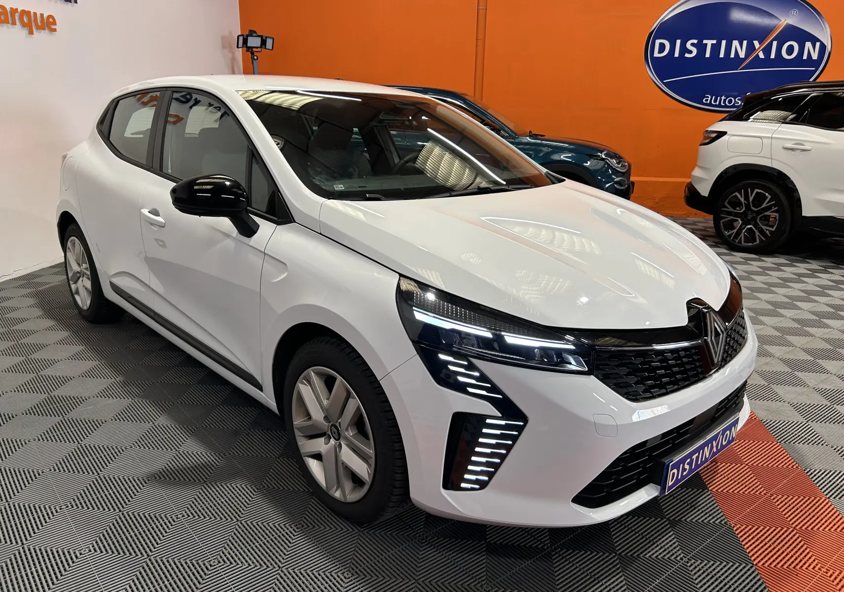 Renault Clio V blanc vue 3/4 avant droit, avec feux LED distinctifs et rétroviseurs noirs dans un showroom.