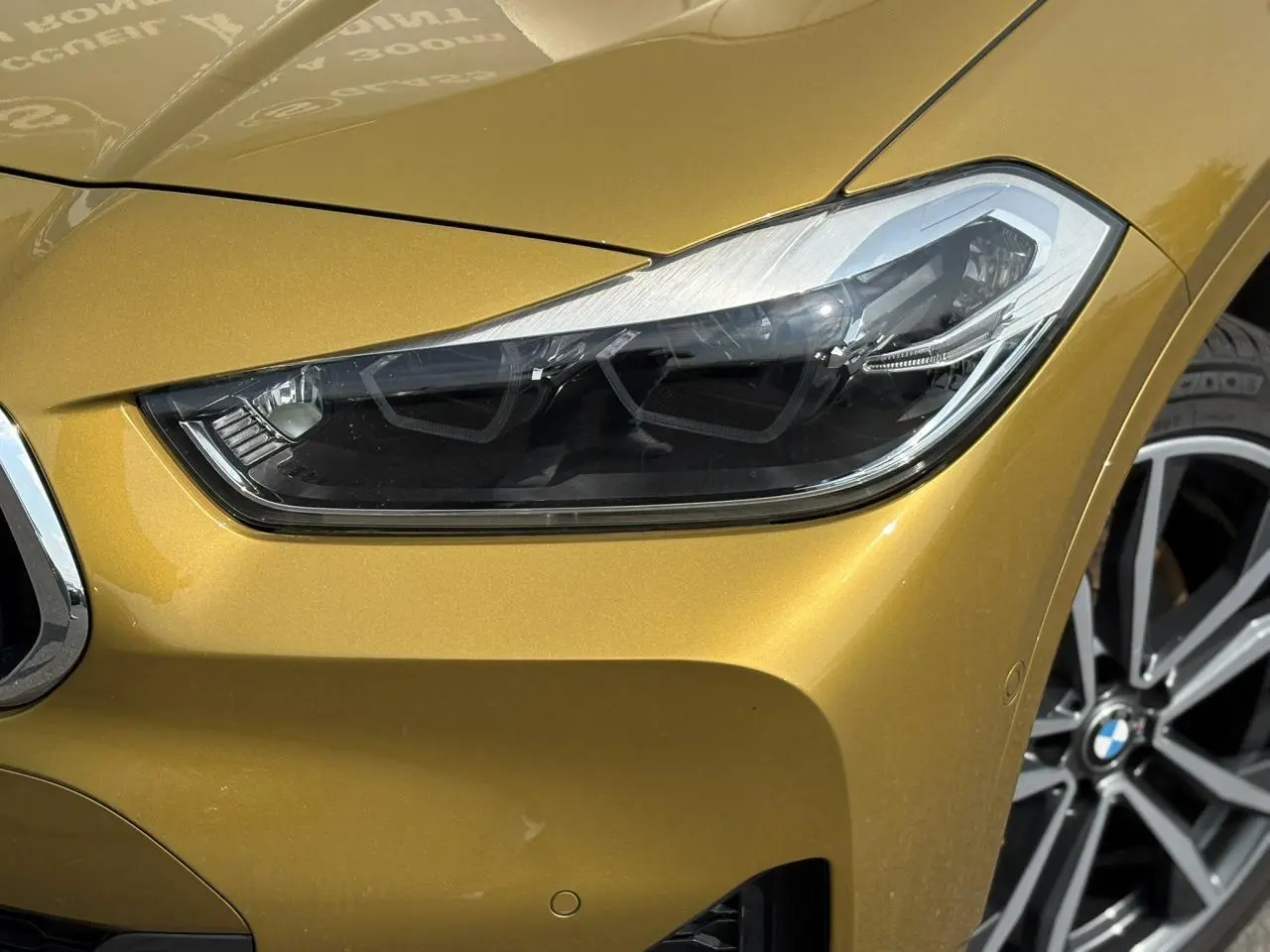 Gros plan sur le phare avant droit et la calandre d'une BMW X2 F39 or Galvanic avec jante alliage bicolore visible.
