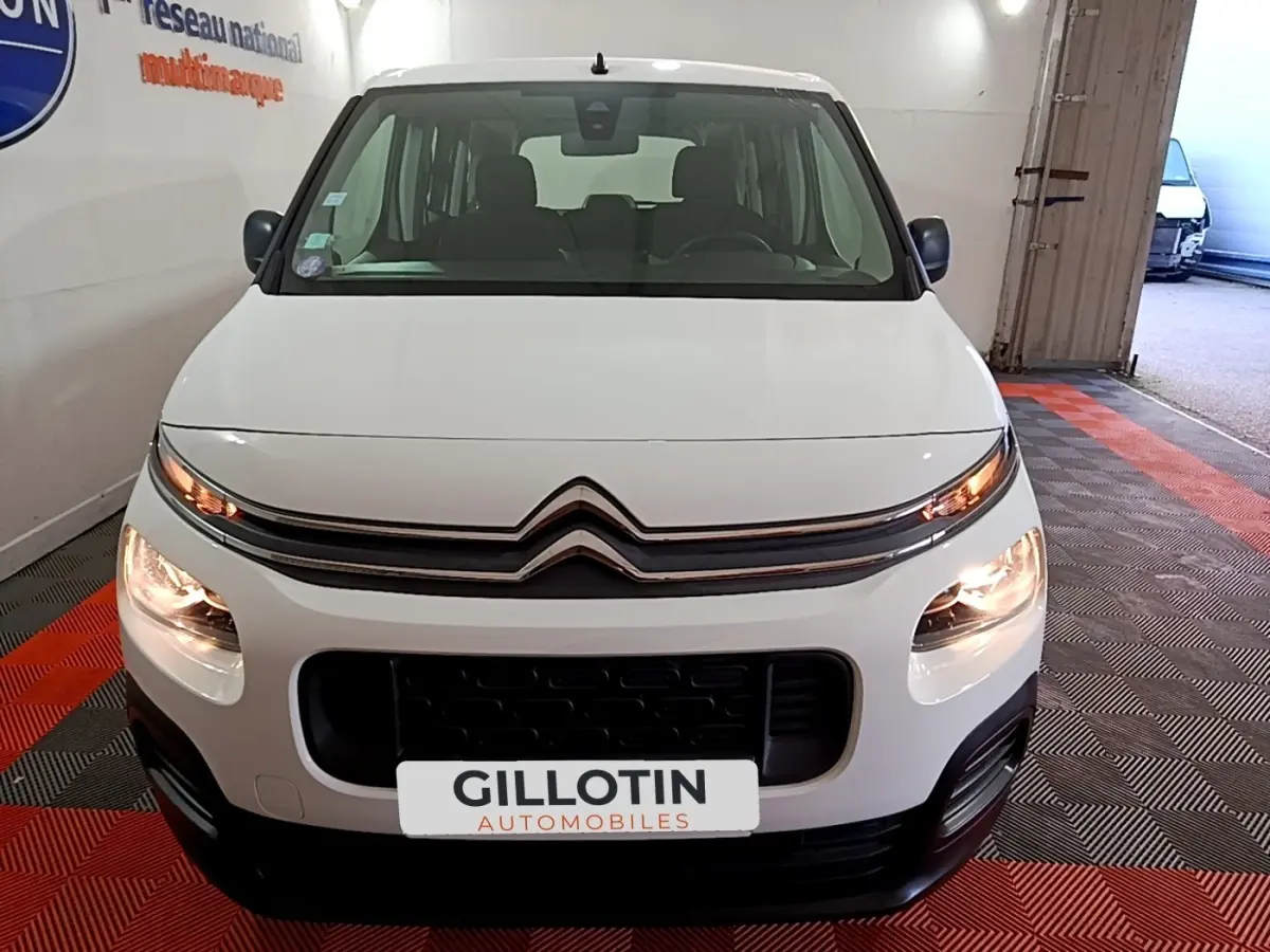 Vue avant d'un Citroën Berlingo XL blanc avec phares allumés, sur sol à damiers rouge et noir en intérieur.