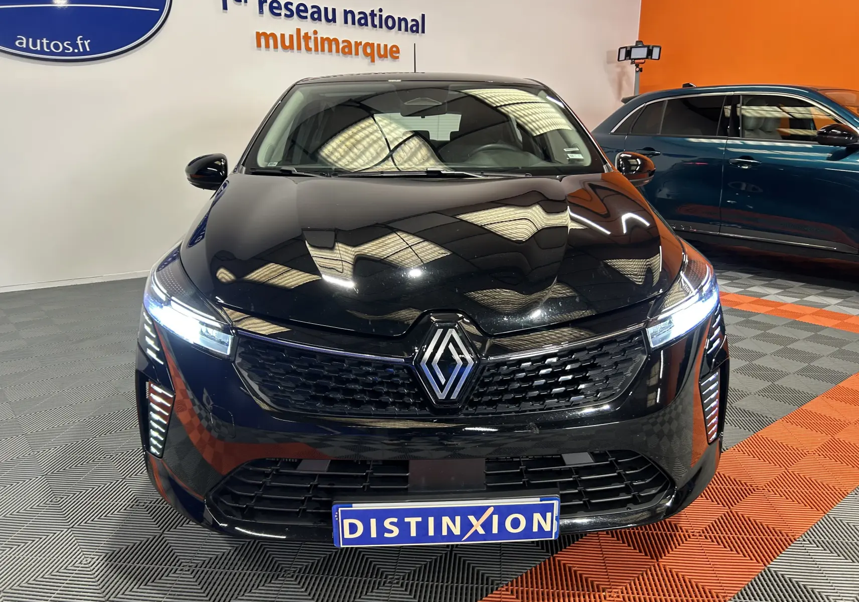 Vue avant d'une Renault Clio V noire avec phares allumés, en intérieur sur sol à damiers gris et orange.