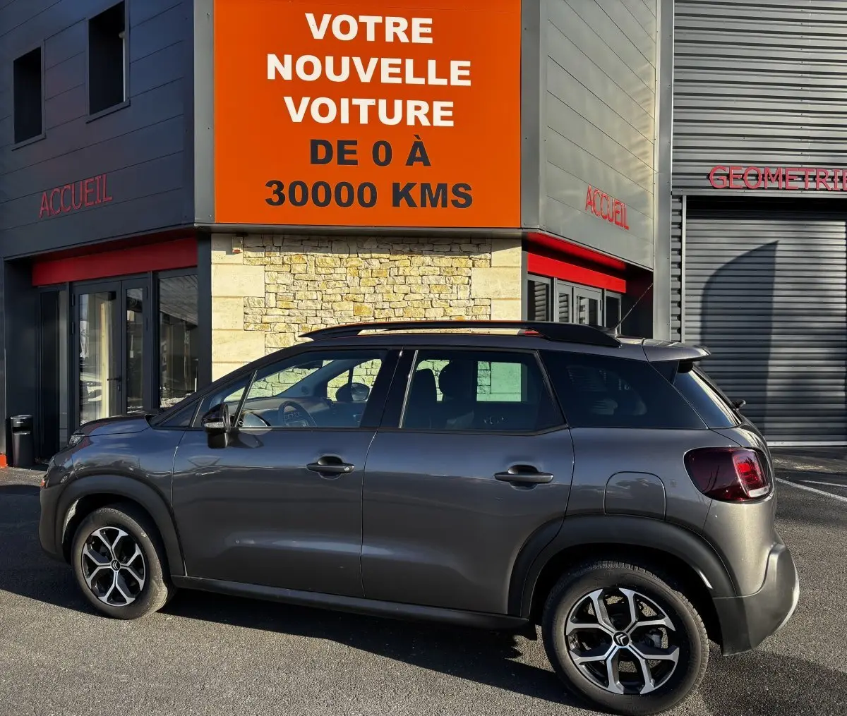 Profil droit d'un Citroën C3 Aircross gris foncé 2024 avec barres de toit noires devant un bâtiment commercial.