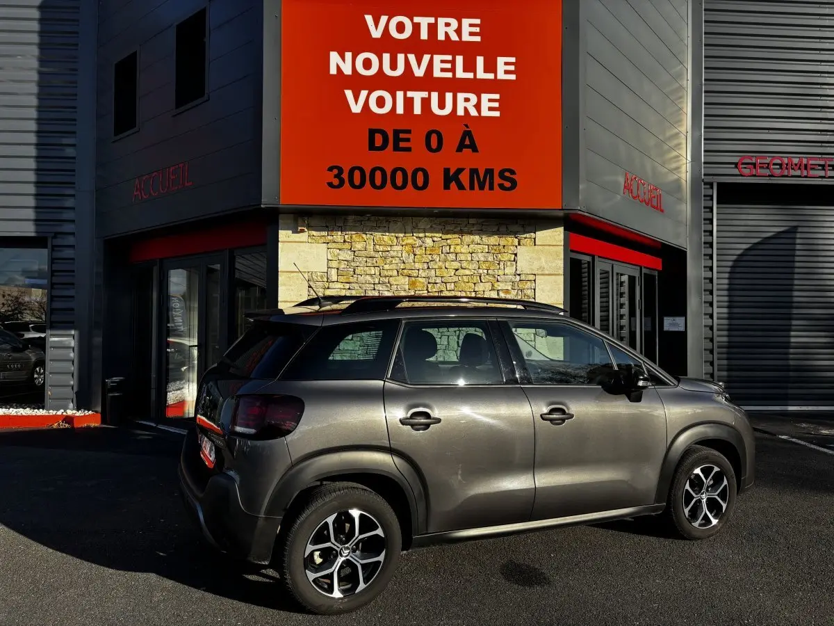 Vue latérale droite d'un Citroën C3 Aircross gris foncé 2024 garé devant un bâtiment avec enseigne rouge.