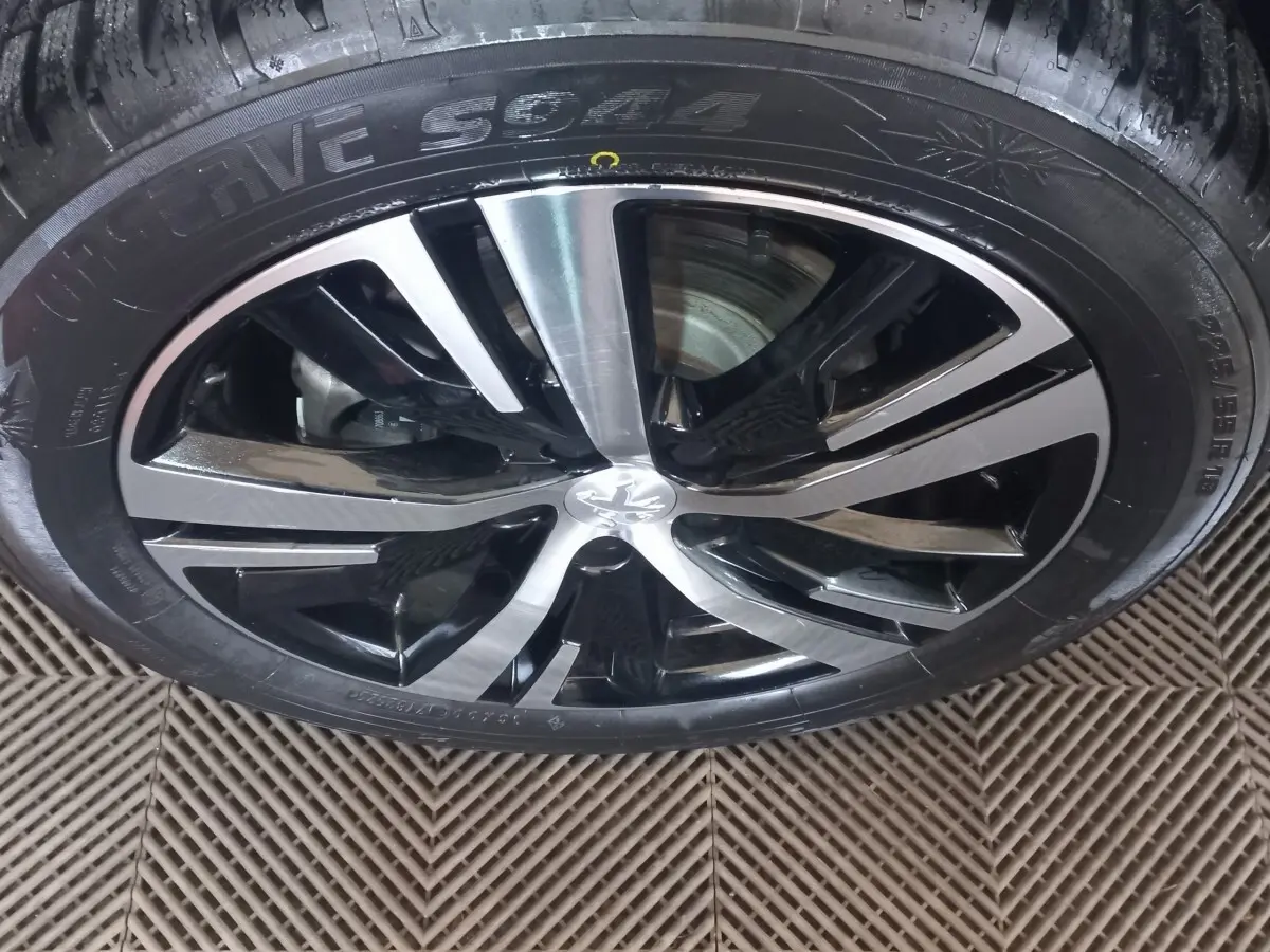 Gros plan sur la jante alliage bi-ton noir et argent avec logo Peugeot, montée sur pneu hiver sur sol texturé.