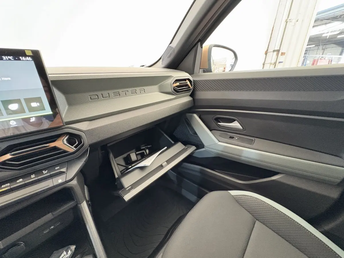 Intérieur du Dacia Duster 2025 côté passager, tableau de bord gris avec boîte à gants ouverte et siège noir.