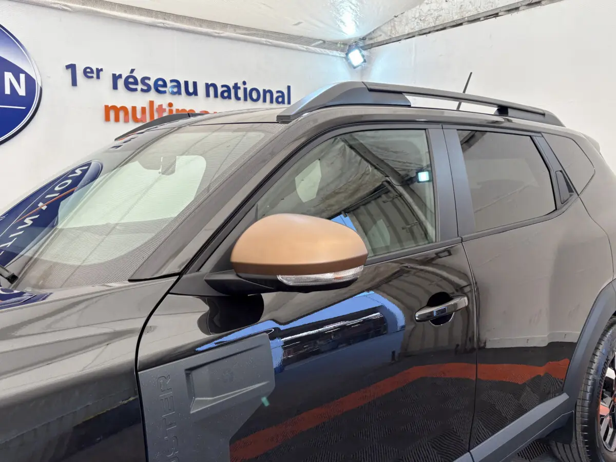 Dacia Duster noir vue côté gauche avant, avec rétroviseur couleur bronze et barres de toit noires.