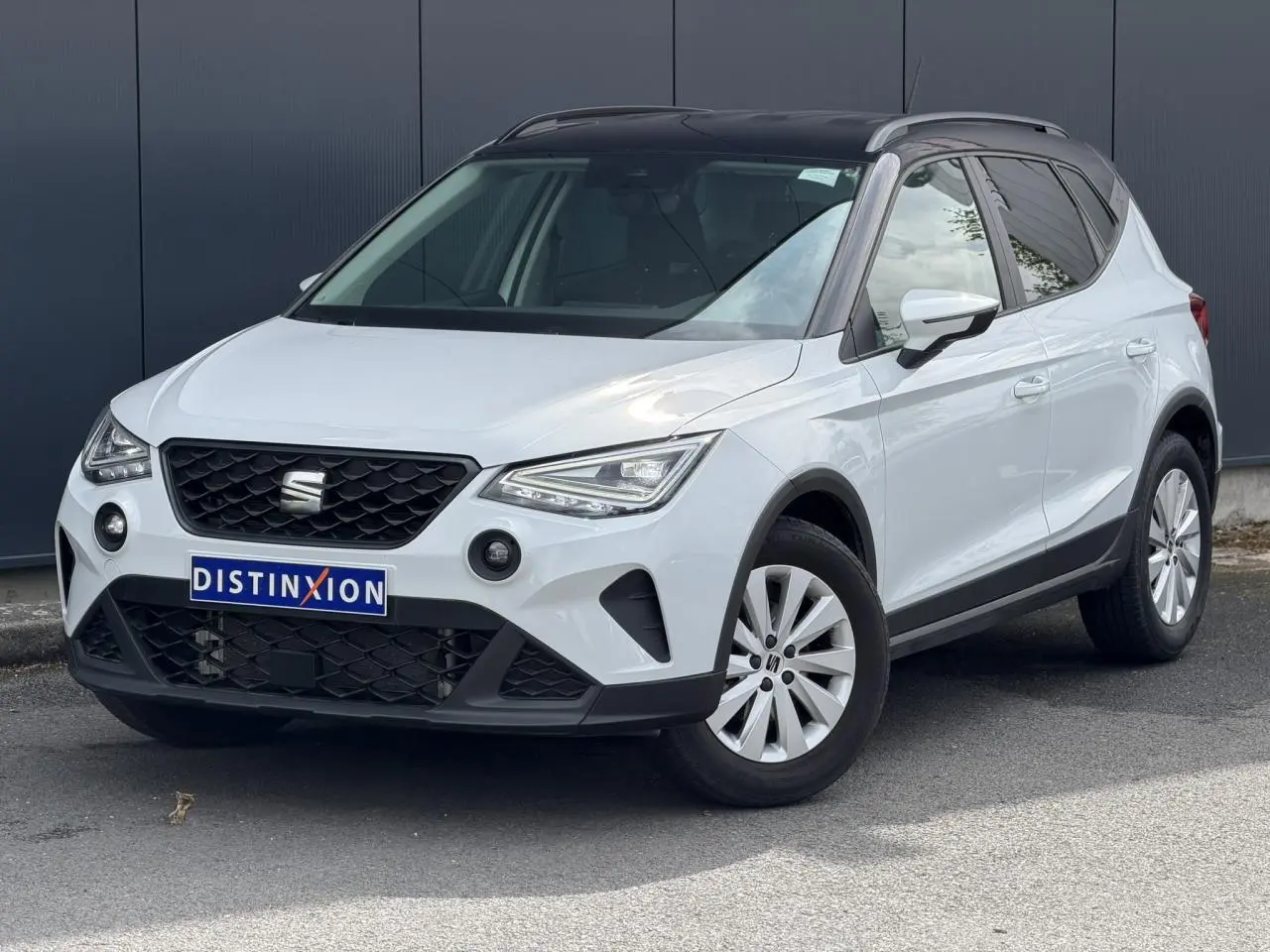 Vue 3/4 avant droite d'un SEAT Arona blanc alpin avec toit noir et jantes alliage 16 pouces, sur fond urbain.