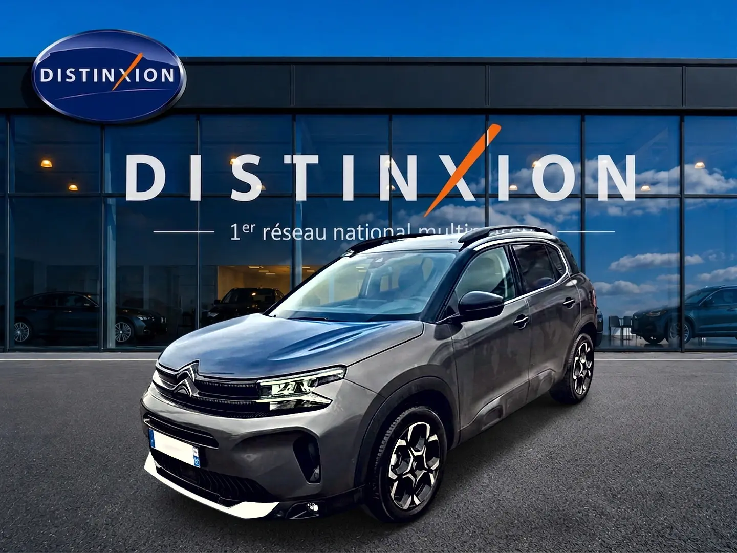 SUV Citroën C5 Aircross gris vu en 3/4 avant droit, avec jantes alu et barres de toit visibles devant un showroom.