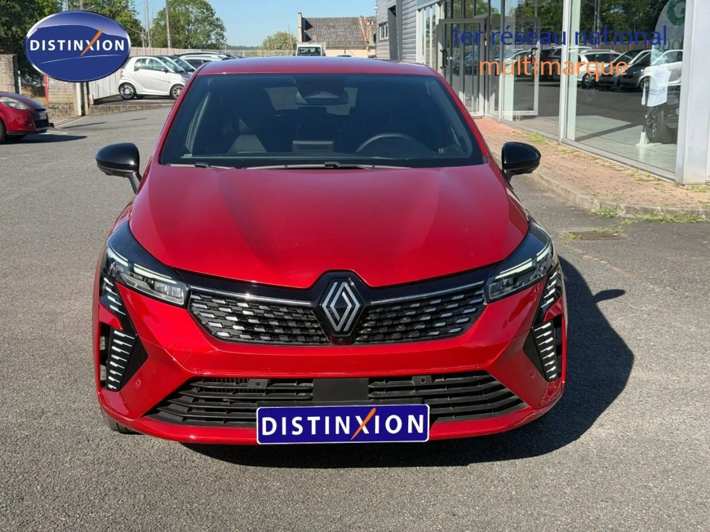 Vue avant d'une Renault Clio rouge avec calandre noire et phares LED, stationnée devant un bâtiment vitré.