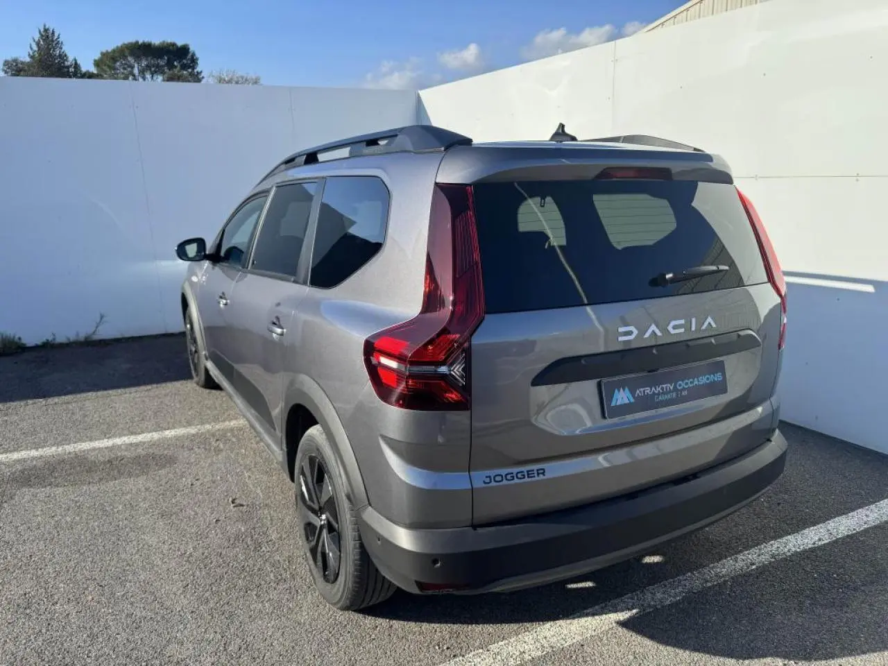 Vue 3/4 arrière gauche d'un Dacia Jogger gris schiste métallisé avec barres de toit et feux arrière LED distinctifs.