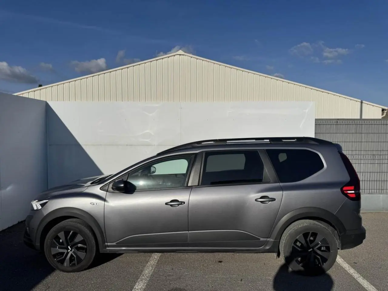 Profil droit du Dacia Jogger gris schiste métallisé avec barres de toit et jantes noires sur parking extérieur.