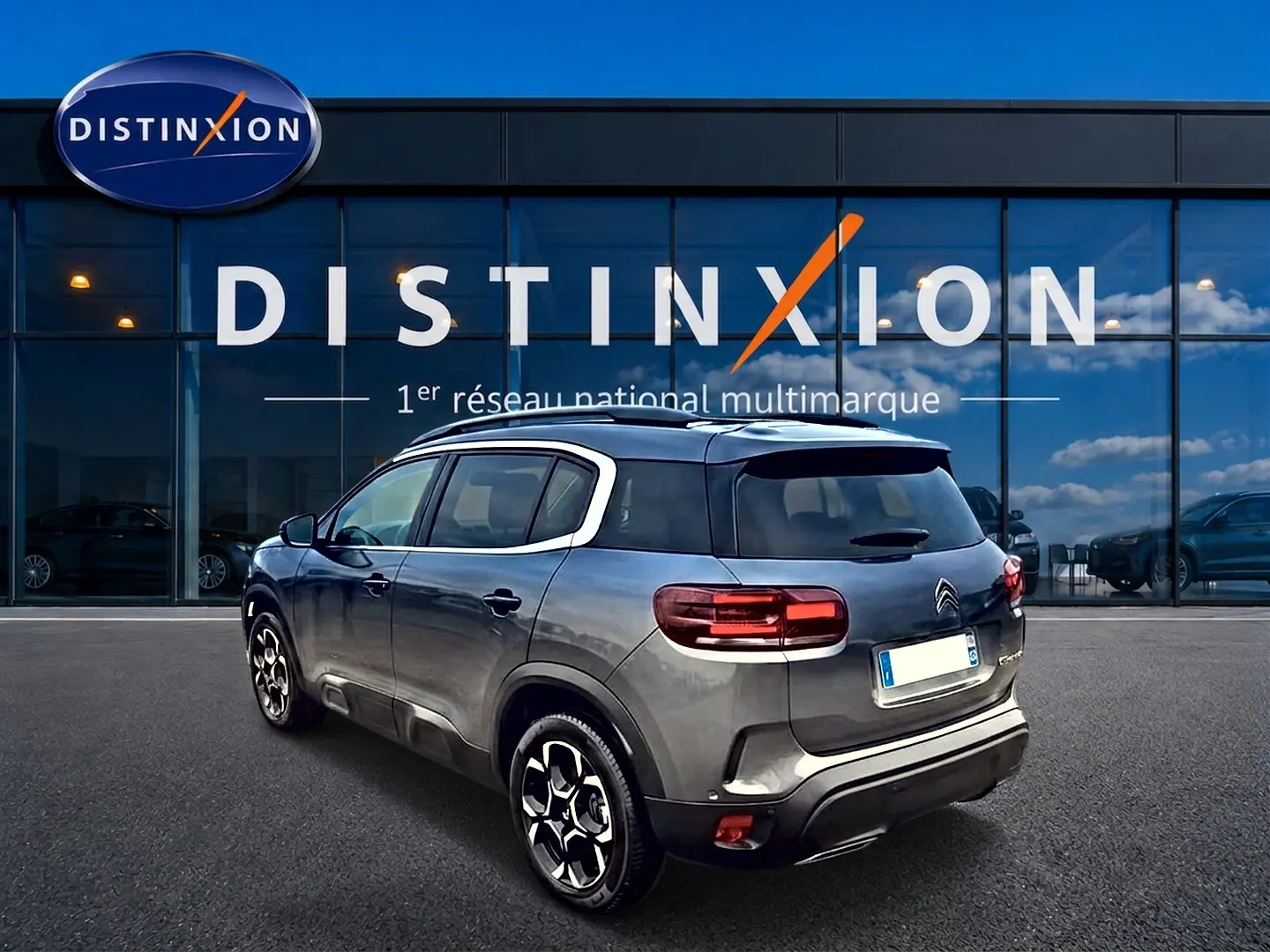 Citroën C5 Aircross gris en 3/4 arrière droit, avec jantes alu et barres de toit visibles devant un showroom Distinxion.