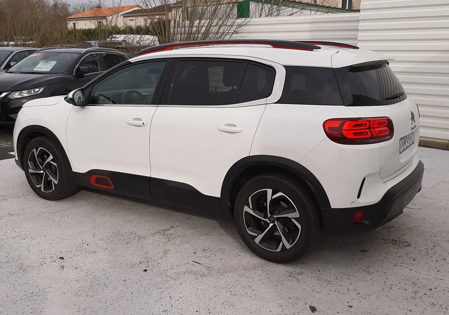 Vue 3/4 arrière droite d'un Citroën C5 Aircross blanc avec toit noir et détails rouges, garé sur un sol clair en extérieur.