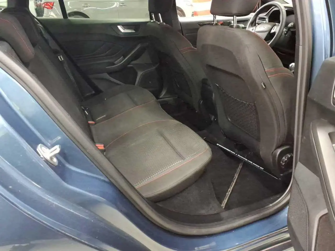 Vue intérieure côté arrière gauche de la Ford Focus SW Bleu Azur, sièges noirs avec surpiqûres rouges et volant visible.