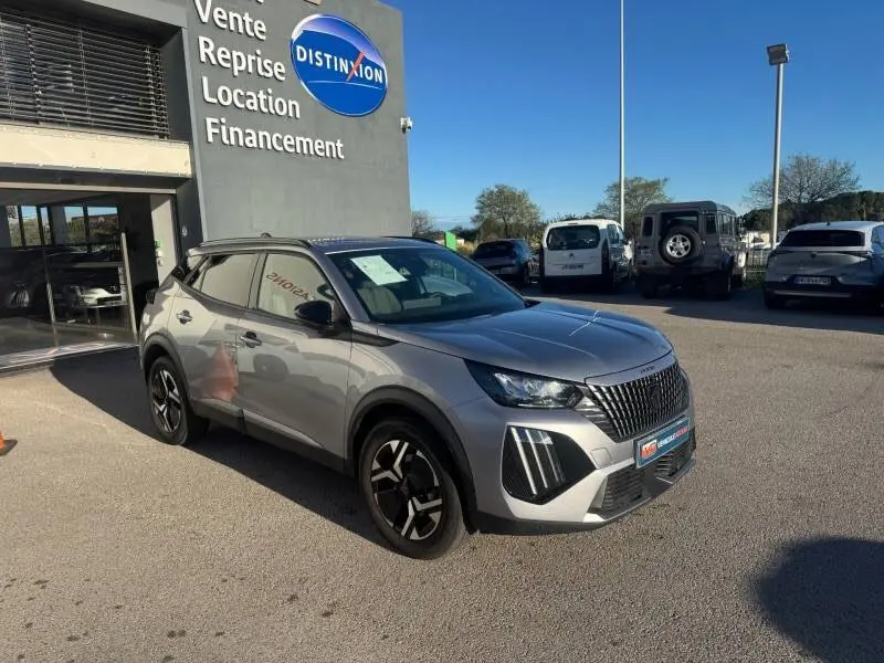 Peugeot 2008 Hybrid gris en 3/4 avant droit avec barres de toit et jantes alliage 17 pouces, devant un concessionnaire.