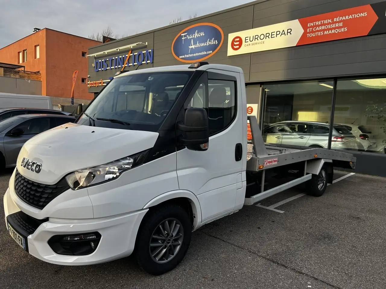 IVECO Daily blanc en 3/4 avant droit, plateau arrière visible, stationné devant un garage avec enseignes.