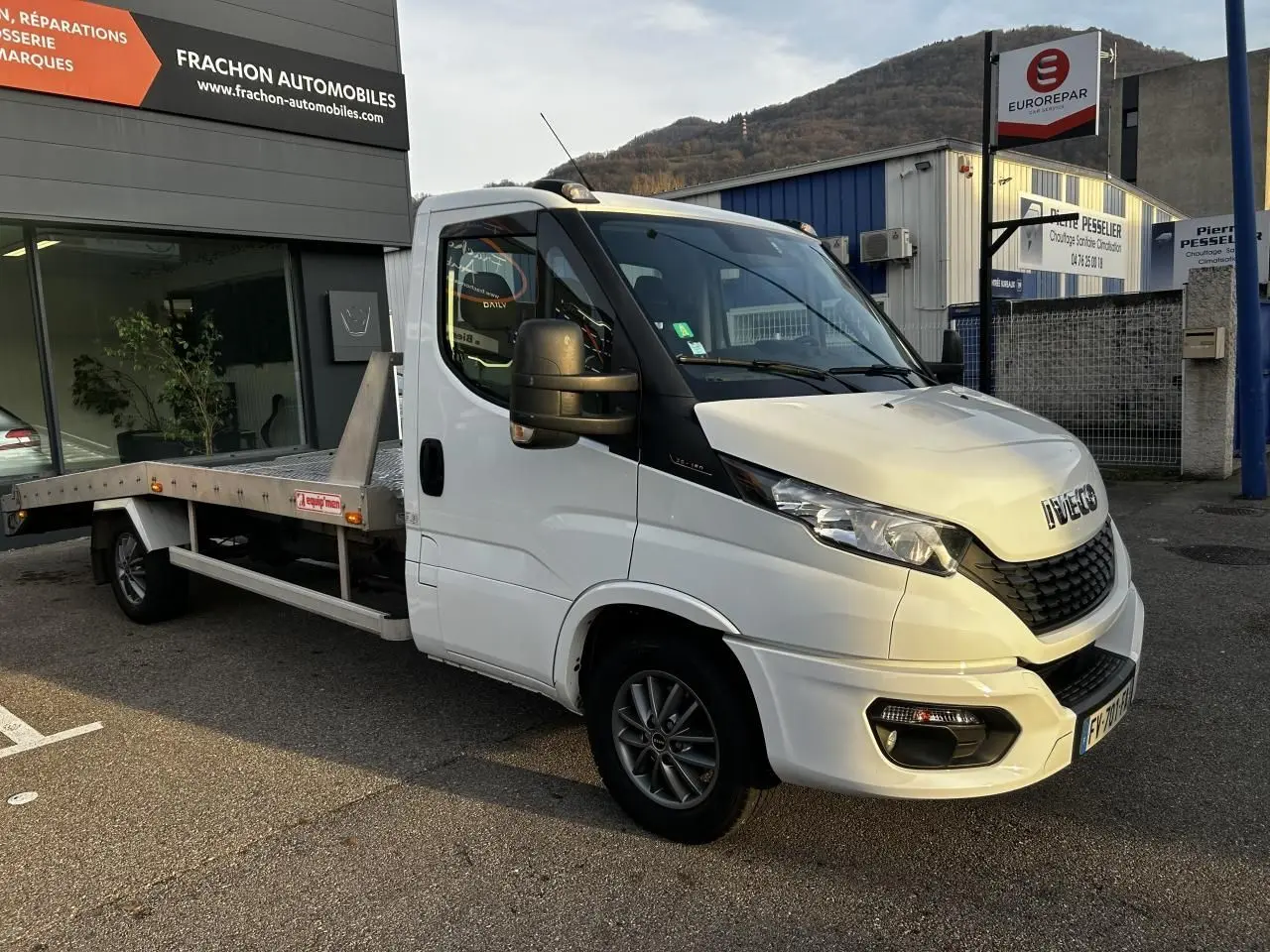 IVECO Daily blanc en 3/4 avant droit, plateau arrière métallique visible, stationné devant un garage automobile.