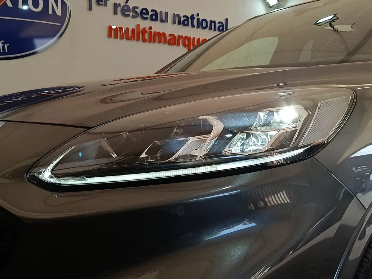 Gros plan sur le phare avant droit allumé du Ford Kuga gris foncé, mettant en valeur les LED et la carrosserie brillante.