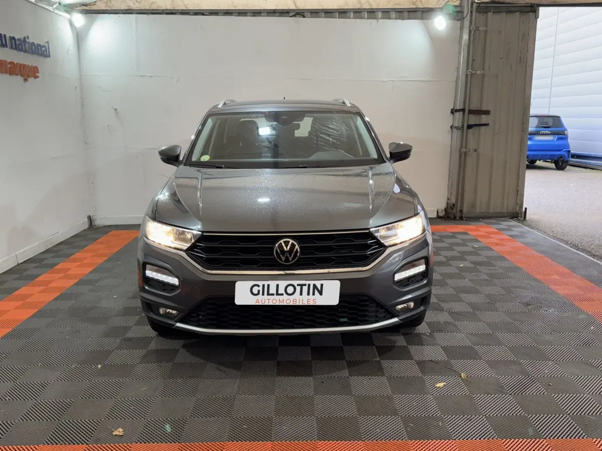 Vue frontale d'un Volkswagen T-Roc gris foncé avec phares allumés et logo VW bien visible sur calandre noire.