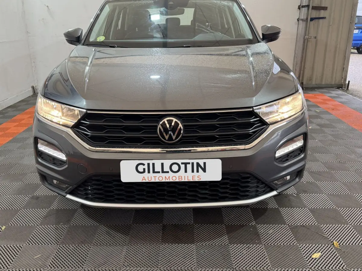 Vue frontale d'un Volkswagen T-Roc gris foncé avec phares allumés et calandre noire hexagonale.
