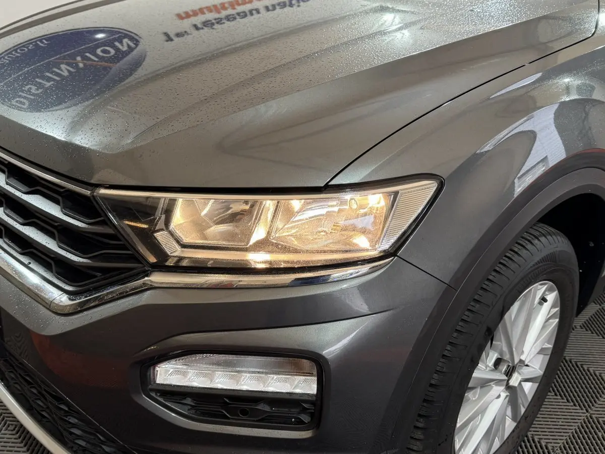 Gros plan sur l'avant droit d'un Volkswagen T-Roc gris foncé avec phares allumés et jante argentée visible.