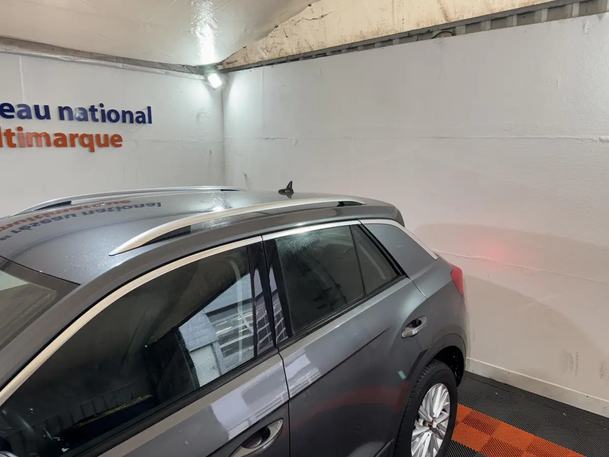 Vue côté gauche arrière du Volkswagen T-Roc gris foncé avec barres de toit argentées dans un garage.