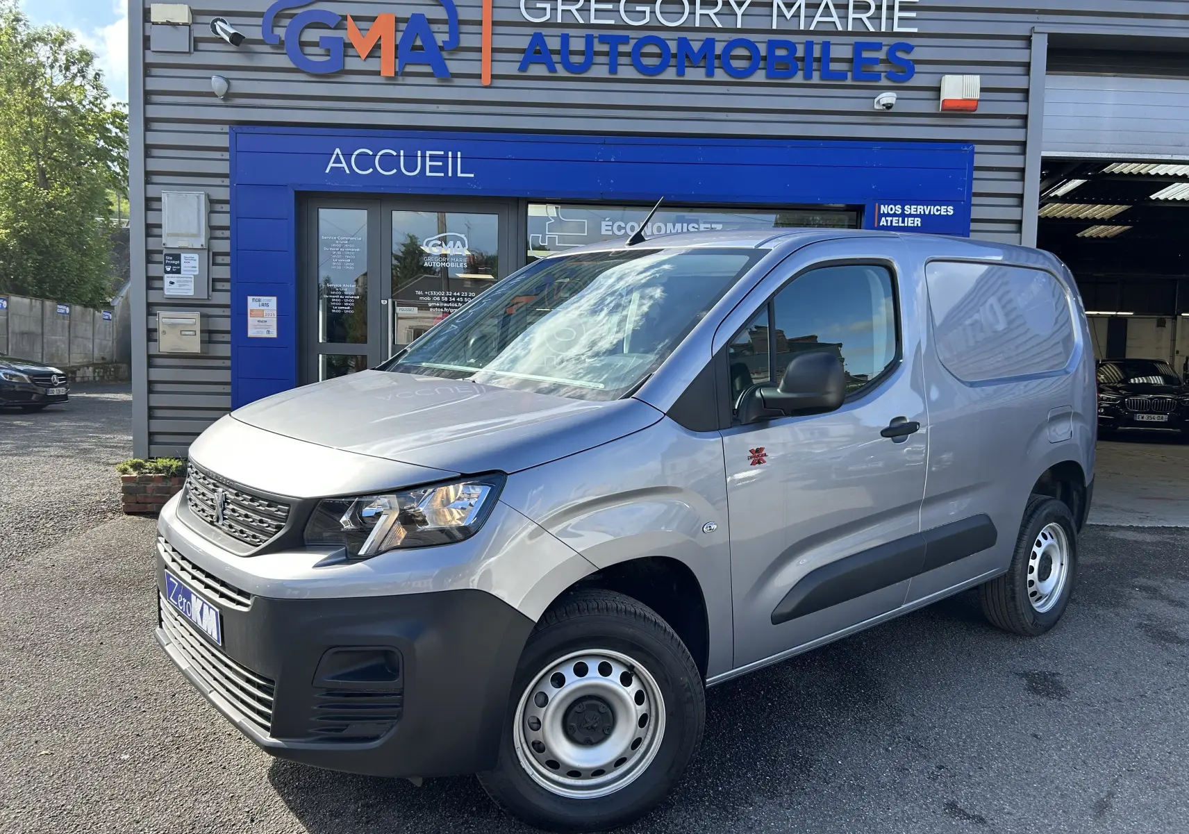 Vue 3/4 avant droit d'un Peugeot Partner Dangel 4x4 gris Artense avec protections noires et jantes acier devant un garage.