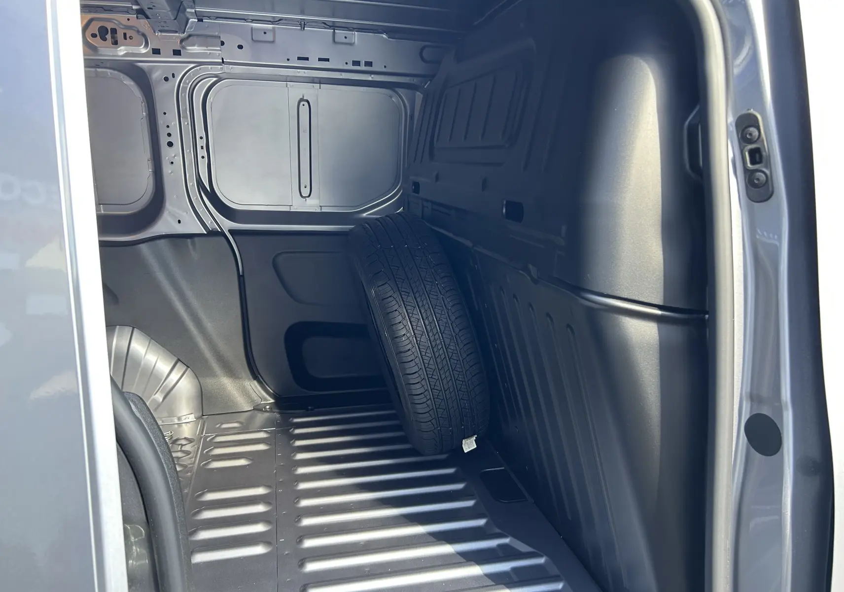 Vue intérieure de la zone de chargement du Peugeot Partner Dangel gris Artense avec une roue de secours appuyée sur la paroi.