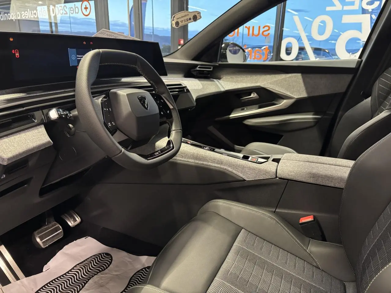 Intérieur avant droit du Peugeot 3008 2025, sellerie tissu gris et cuir, planche de bord avec décor tissu chiné et volant compact noir.