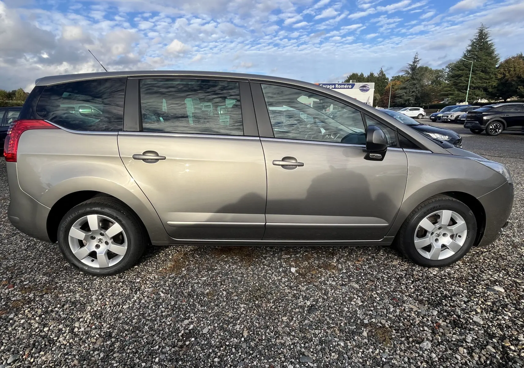 Vue latérale côté gauche d'un Peugeot 5008 gris métallisé de 2013 garé sur un sol gravillonné.