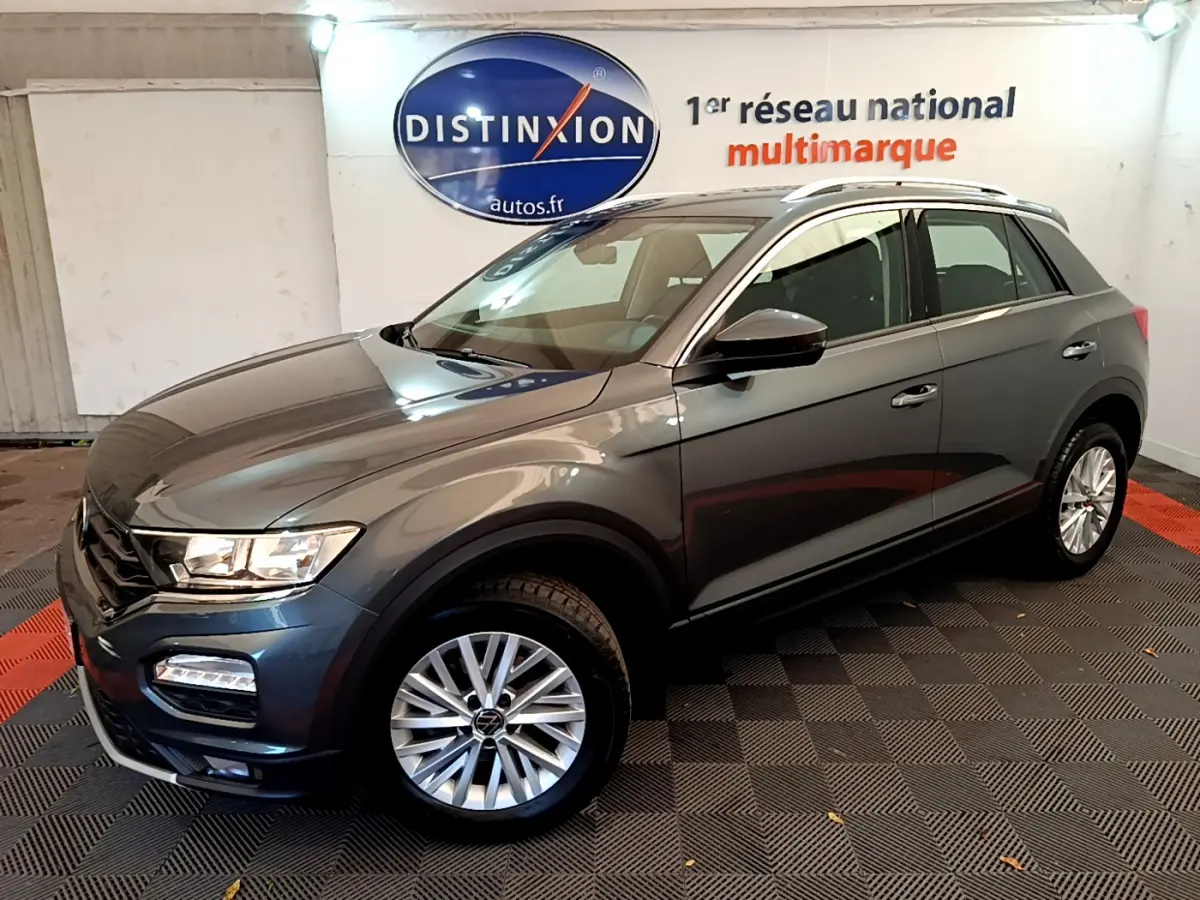 Volkswagen T-Roc gris foncé vue 3/4 avant droit, avec jantes alliage et barres de toit argentées.