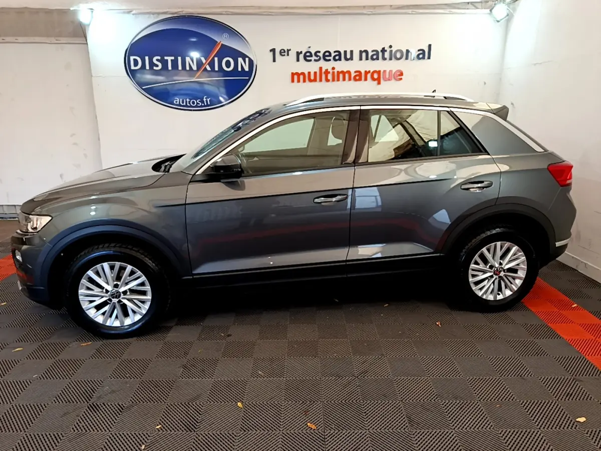 Profil côté gauche d'un Volkswagen T-Roc gris foncé 2021 avec jantes alliage et rails de toit blancs dans un showroom.
