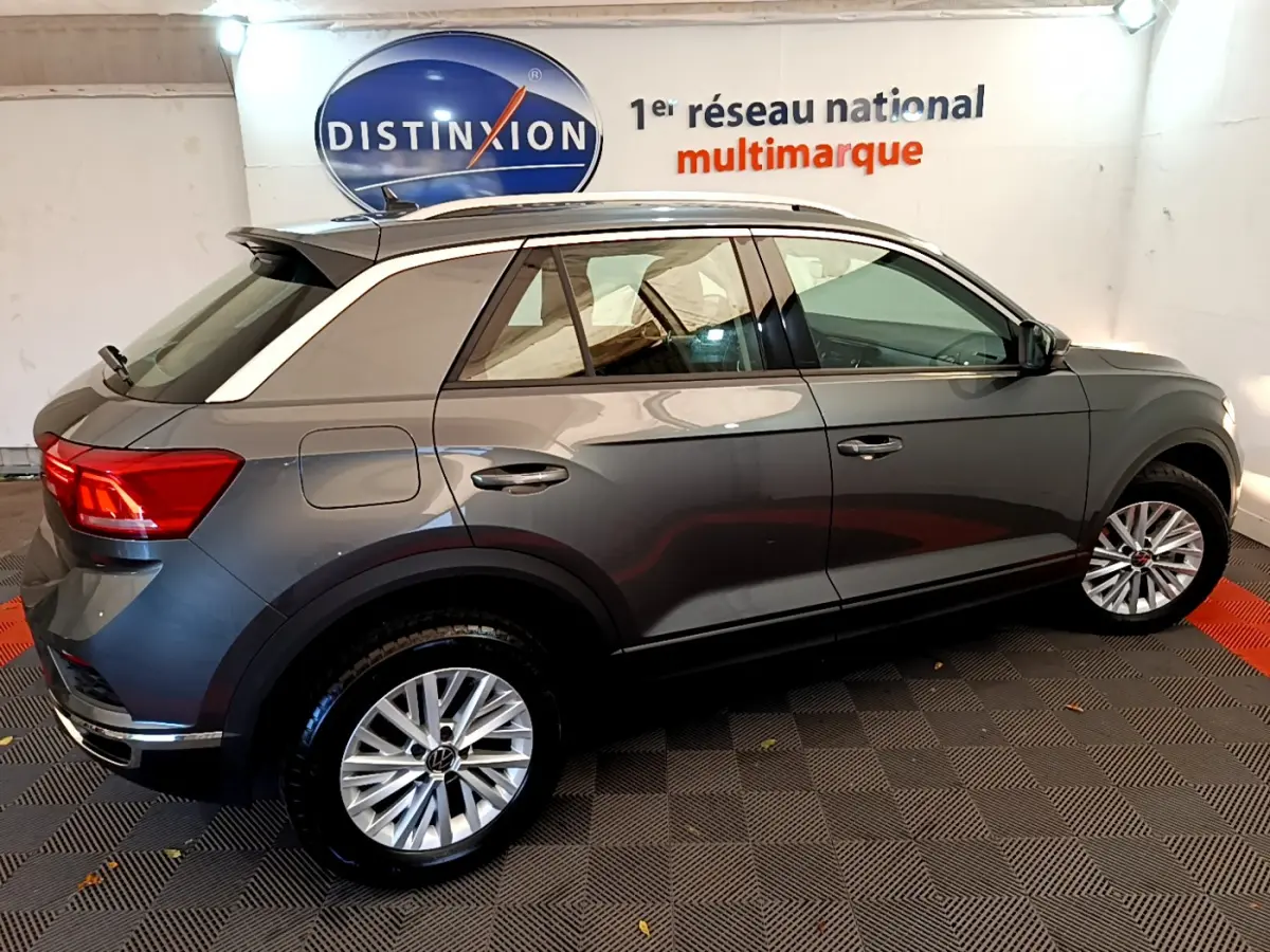 Vue de profil côté droit d'un Volkswagen T-Roc gris foncé avec jantes alliage et rails de toit blancs dans un showroom.