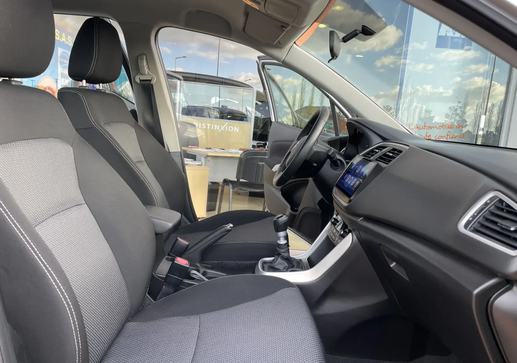 Intérieur du Suzuki SX4 S-Cross 2016 vu côté conducteur, sièges noirs, tableau de bord avec écran tactile et levier de vitesses manuel.