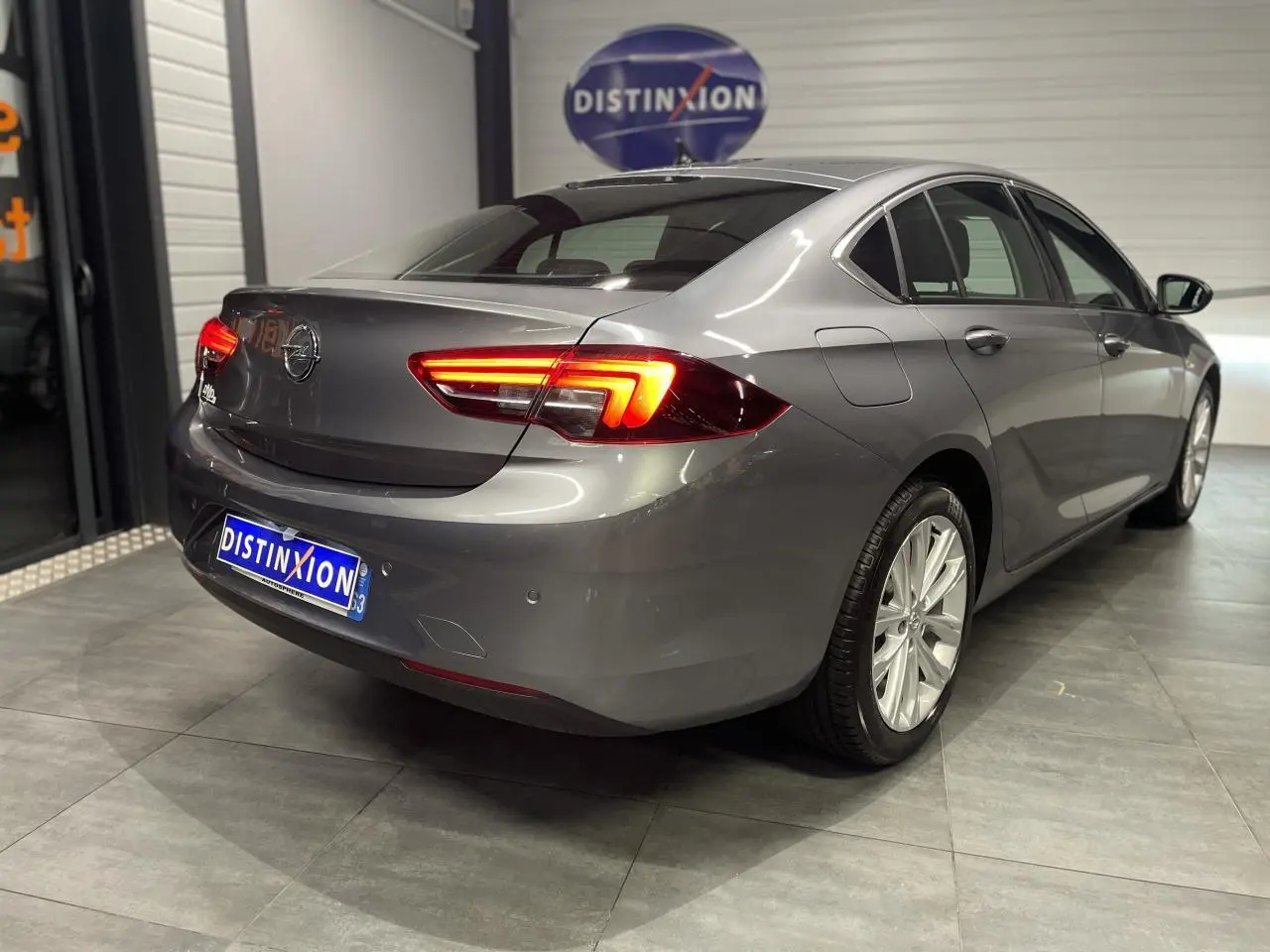 Opel Insignia Grand Sport gris clair vue 3/4 arrière droit avec feux arrière LED allumés en intérieur showroom.