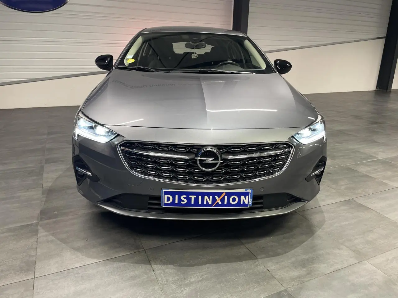 Opel Insignia Grand Sport gris clair vue de face avec calandre noire et phares LED allumés en intérieur showroom.