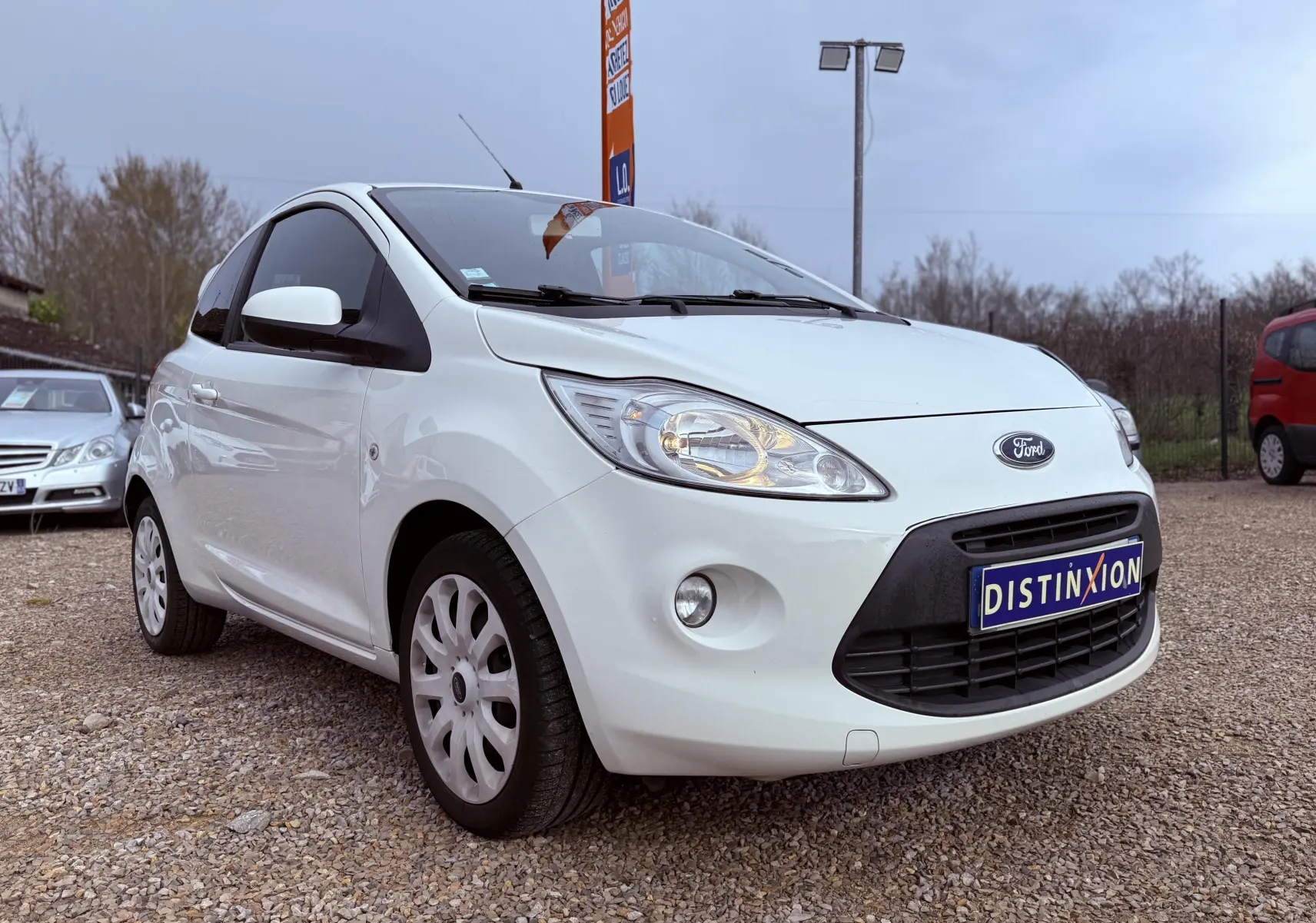 Ford Ka blanc vue 3/4 avant droit sur parking gravillonné avec plaque Distinxion visible.