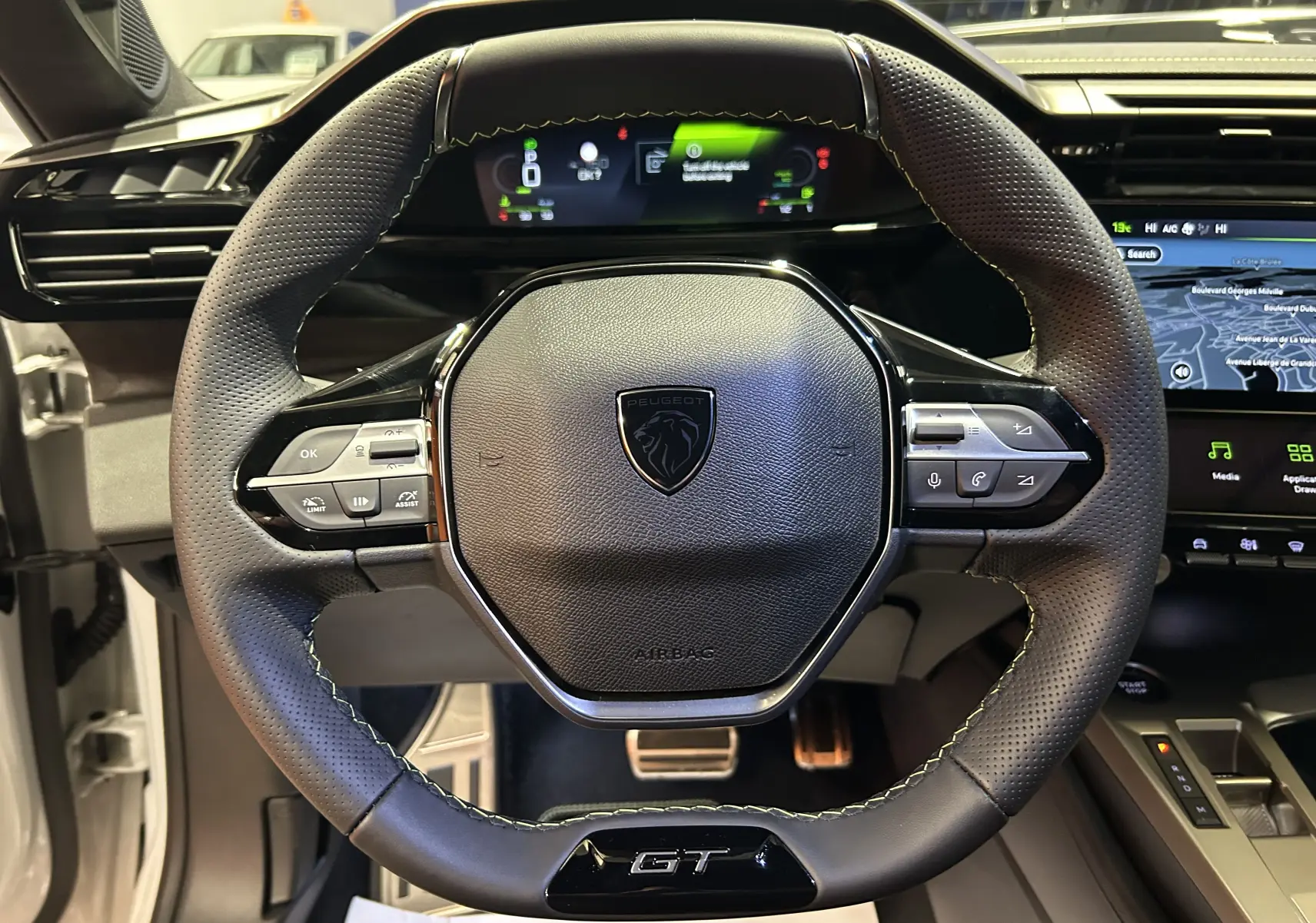 Vue rapprochée du volant cuir perforé noir avec logo Peugeot et badge GT, tableau de bord numérique et écran GPS visibles.