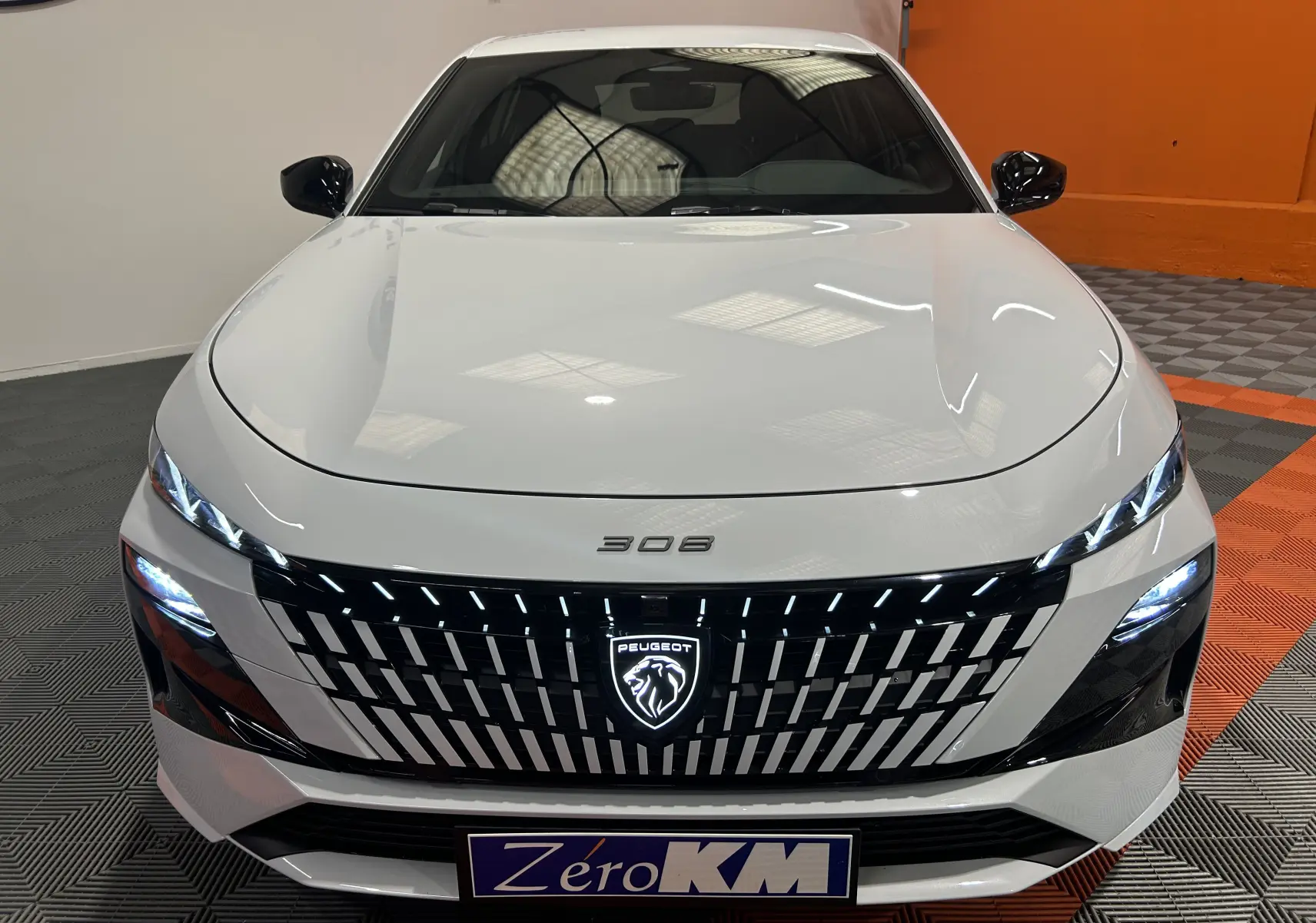 Vue frontale d’une Peugeot 308 blanche Okenite avec calandre noire et logo lumineux au centre.