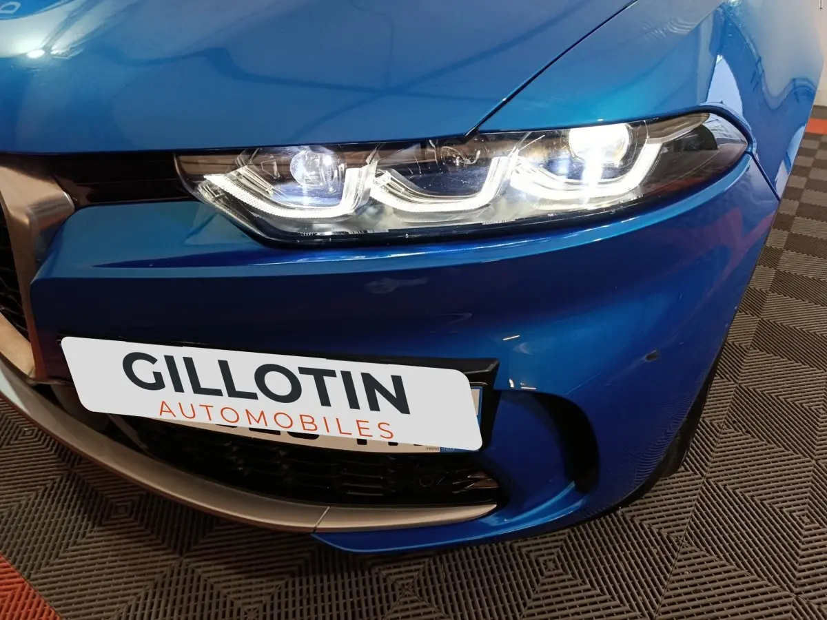 Gros plan sur le phare avant droit allumé d'une Alfa Romeo Tonale bleu 2023 avec calandre et plaque avant visibles.